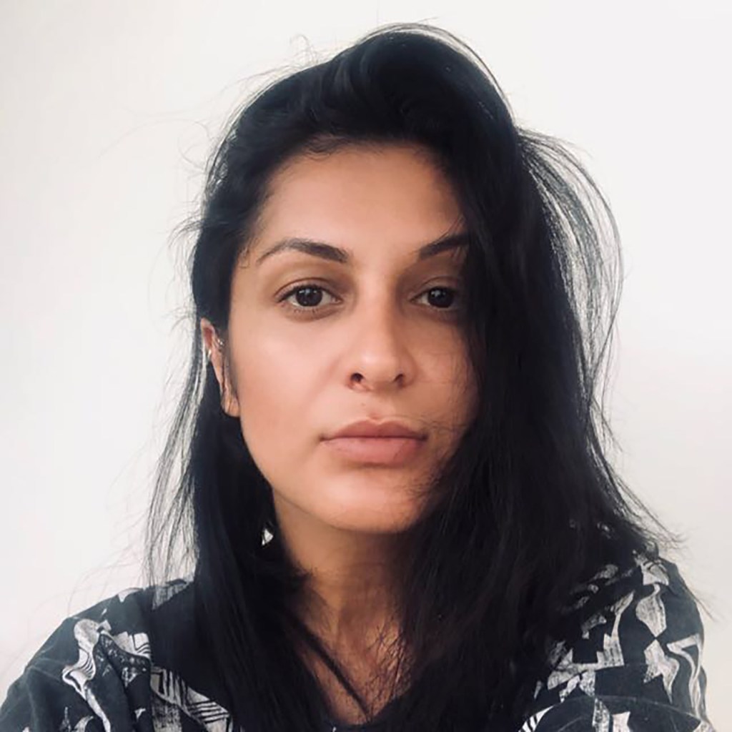 A close-up portrait of a woman with long dark hair, wearing a patterned shirt, against a plain background.
